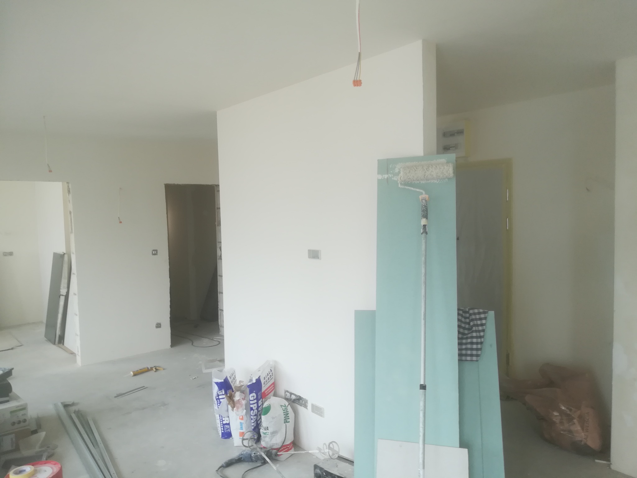 Wnętrze w trakcie remontu: widoczne płyty kartonowo-gipsowe o błękitnym odcieniu oparte o ścianę, wałek malarski, worki z gipsem, mikser budowlany i narzędzia na podłodze, surowe ściany...