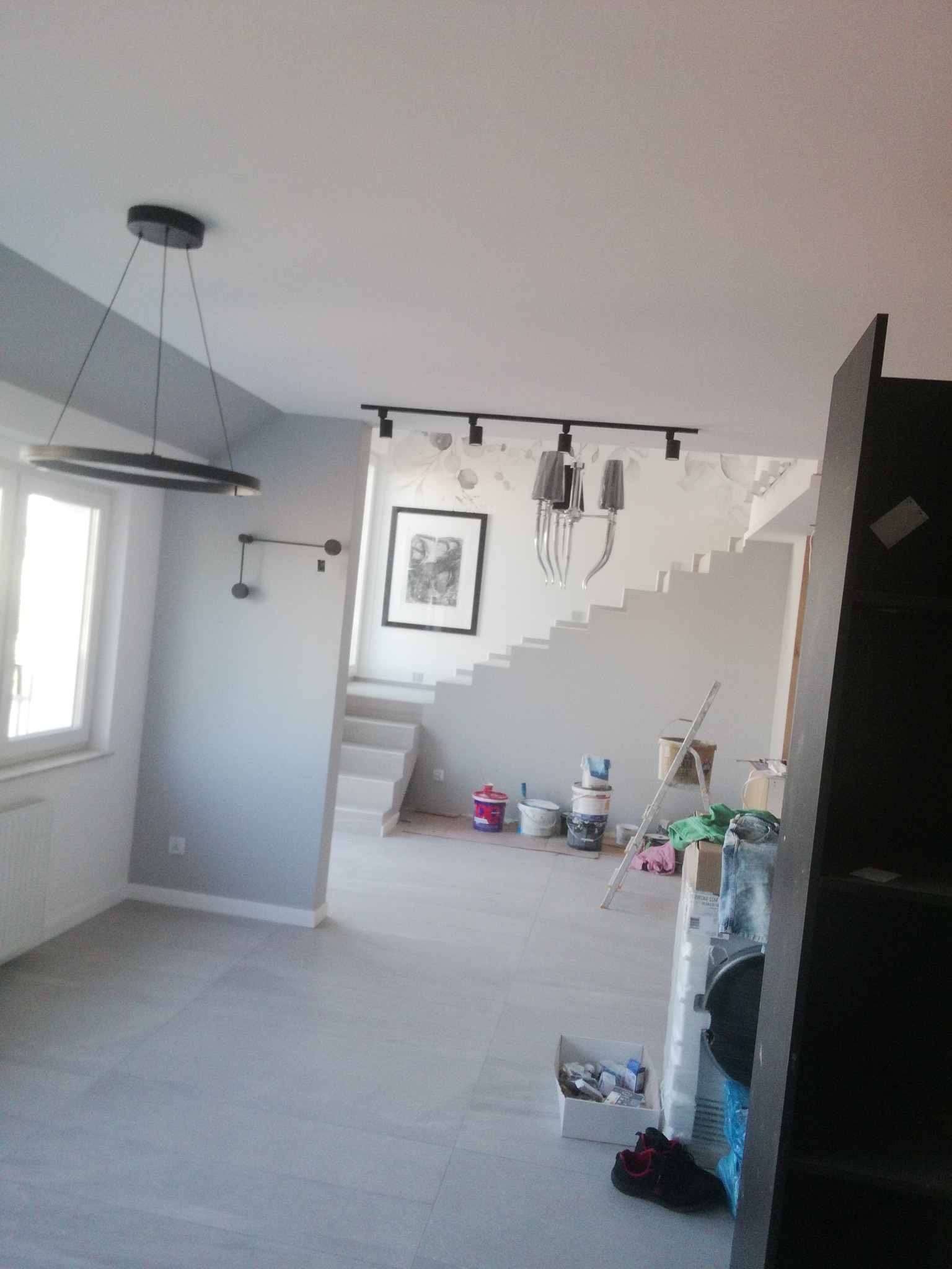 Wnętrze domu w trakcie remontu, widoczne świeżo pomalowane szare ściany, białe schody, nowoczesne lampy sufitowe i akcesoria malarskie, takie jak pędzle i farby.