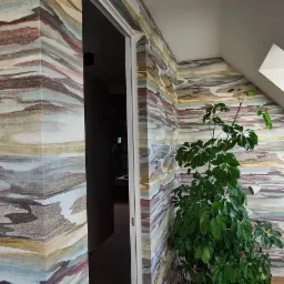 Wnętrze poddasza z wzorzystą tapetą imitującą krajobraz, widoczne okno dachowe i roślina doniczkowa.