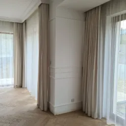Jasne, eleganckie zasłony w minimalistycznym wnętrzu z parkietem ułożonym w jodełkę. Zasłony zawieszone na ukrytych karniszach szynowych, wpuszczonych we wnękę sufitową. Widoczny fragment okna...