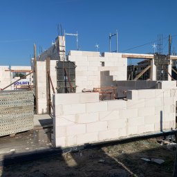 GREAT HOUSE Marcin Kobylarz - Wznoszenie ścian domu z betonu komórkowego Solbet. Widoczne szalunki, zbrojenia i ułożone bloczki. Jasne niebo w tle. Etap budowy w słoneczny dzień.