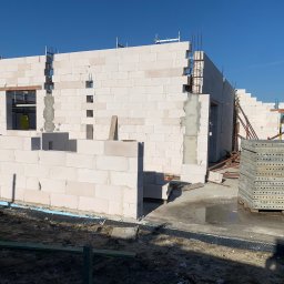 GREAT HOUSE Marcin Kobylarz - Wznoszenie ścian domu z betonu komórkowego. Widoczne zbrojenia i otwory okienne. Na pierwszym planie stos szalunków. Budowa w słoneczny dzień.