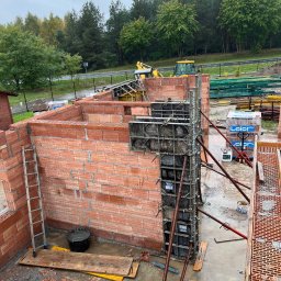 GREAT HOUSE Marcin Kobylarz - Wznoszenie ścian domu z czerwonej cegły, widoczne szalunki i zbrojenia. W tle drzewa i sprzęt budowlany. Ujęcie z góry, ukazujące postęp prac murarskich.