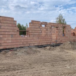 GREAT HOUSE Marcin Kobylarz - Wznoszone ściany domu z czerwonej cegły ceramicznej, widoczne otwory okienne i drzwiowe, teren budowy z piaskiem i narzędziami, błękitne niebo w tle.