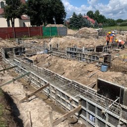 GREAT HOUSE Marcin Kobylarz - Zbrojenie fundamentów pod budowę domu jednorodzinnego. Widoczne szalunki, stalowe pręty i pracownicy w odblaskowych kamizelkach. W tle budynek i drzewa.