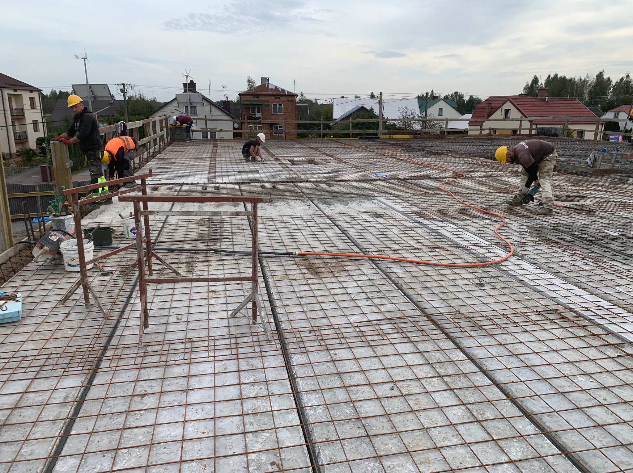 Zbrojenie stropu domu jednorodzinnego z widocznymi pracownikami w kaskach, przygotowującymi konstrukcję pod wylanie betonu. Widok z góry na plac budowy.
