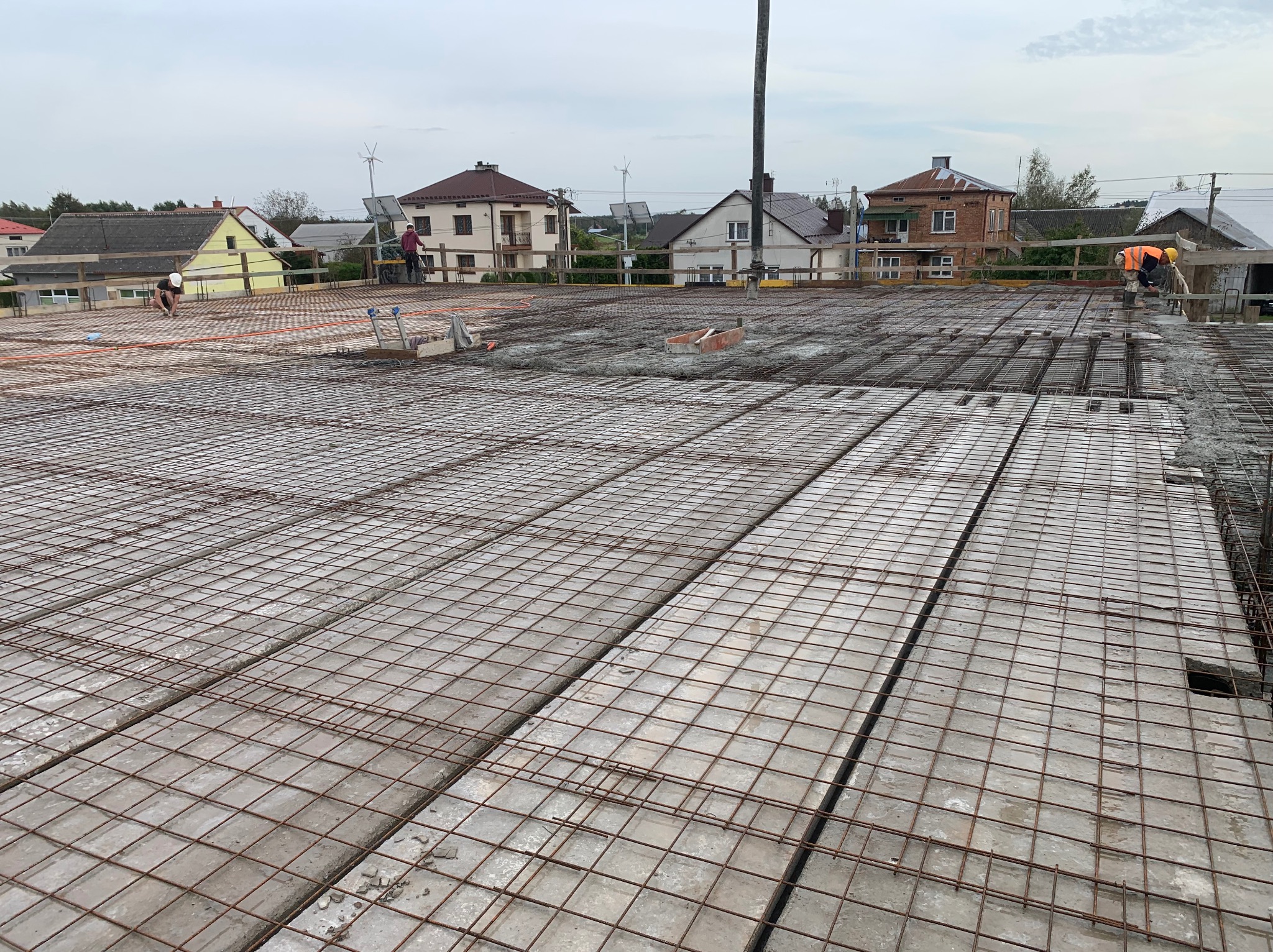 Zbrojenie stropu Teriva z betonem, widoczne płyty stropowe i siatka zbrojeniowa. W tle domy jednorodzinne i pracownicy budowlani w kaskach.