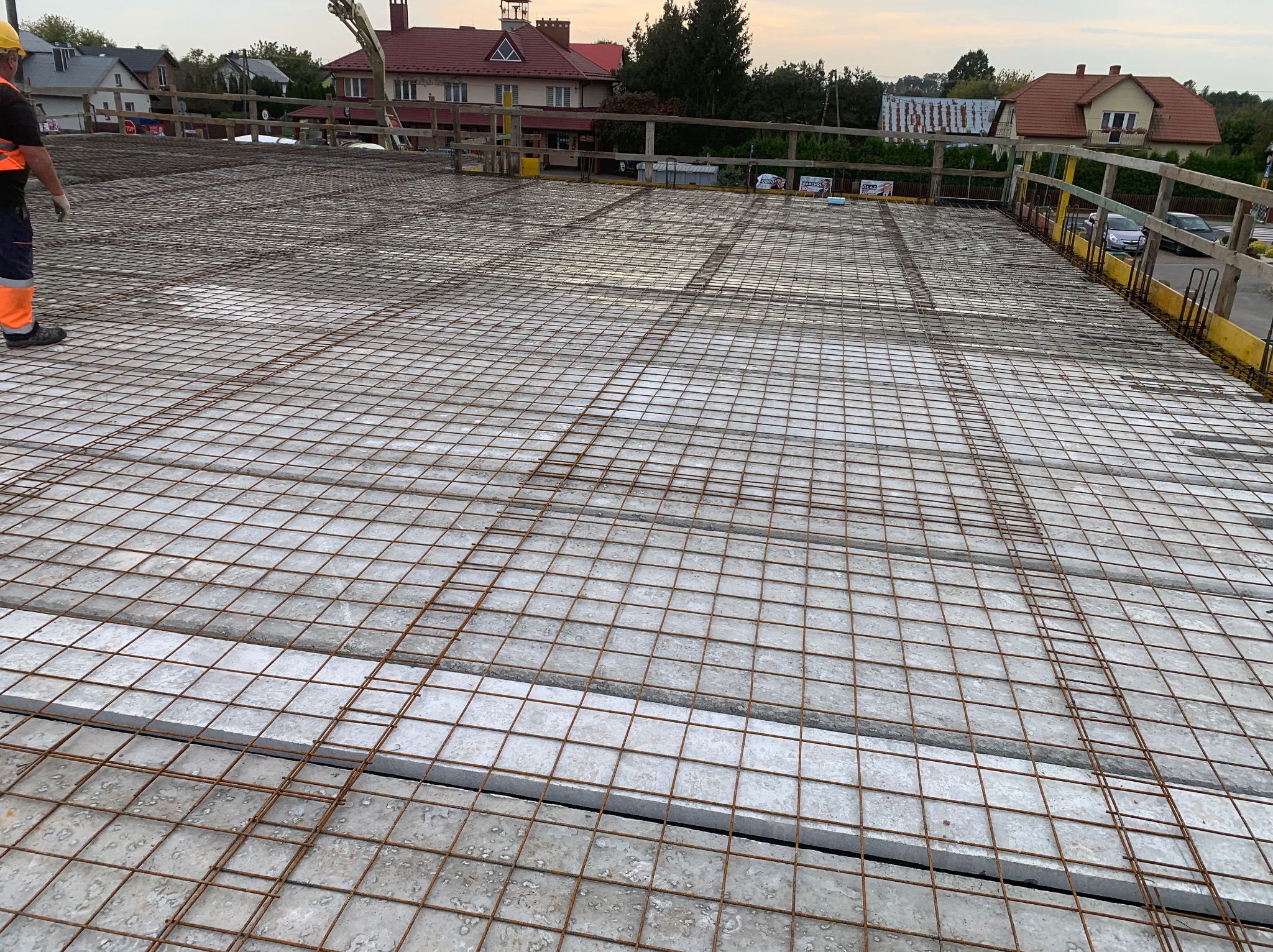 Siatka zbrojeniowa na stropie budynku w trakcie budowy, widok z góry. W tle zabudowa jednorodzinna i pochmurne niebo.
