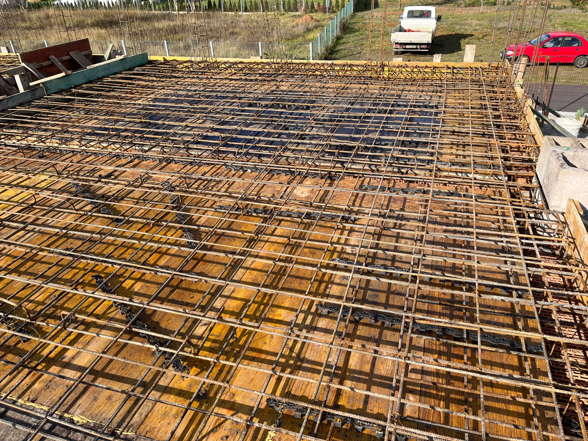 Zbrojenie płyty fundamentowej na deskowaniu, widok z góry. Na drugim planie samochody i zieleń. Słoneczny dzień.