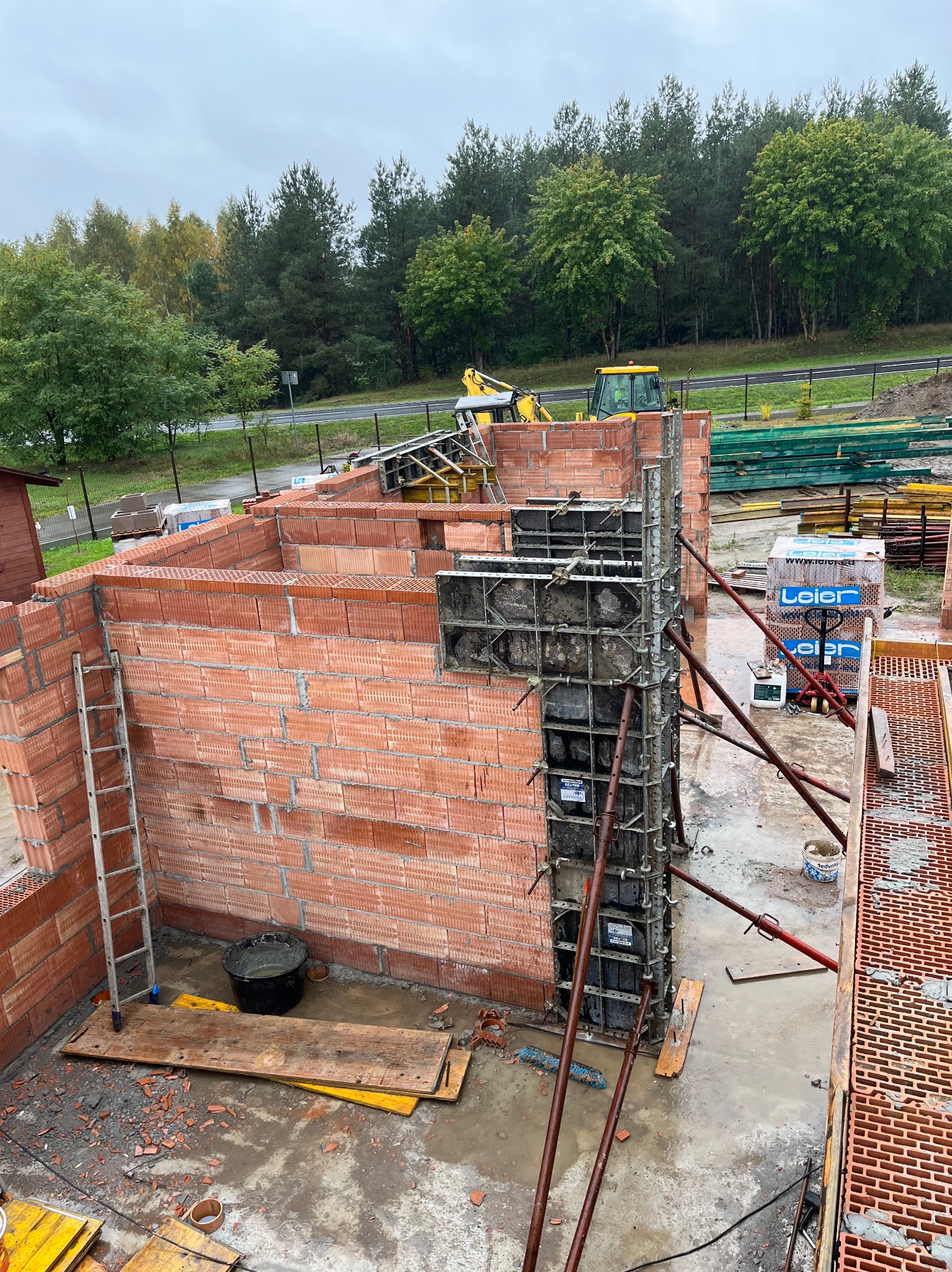 Wznoszenie ścian domu z czerwonej cegły, widoczne szalunki i zbrojenia. W tle drzewa i sprzęt budowlany. Ujęcie z góry, ukazujące postęp prac murarskich.
