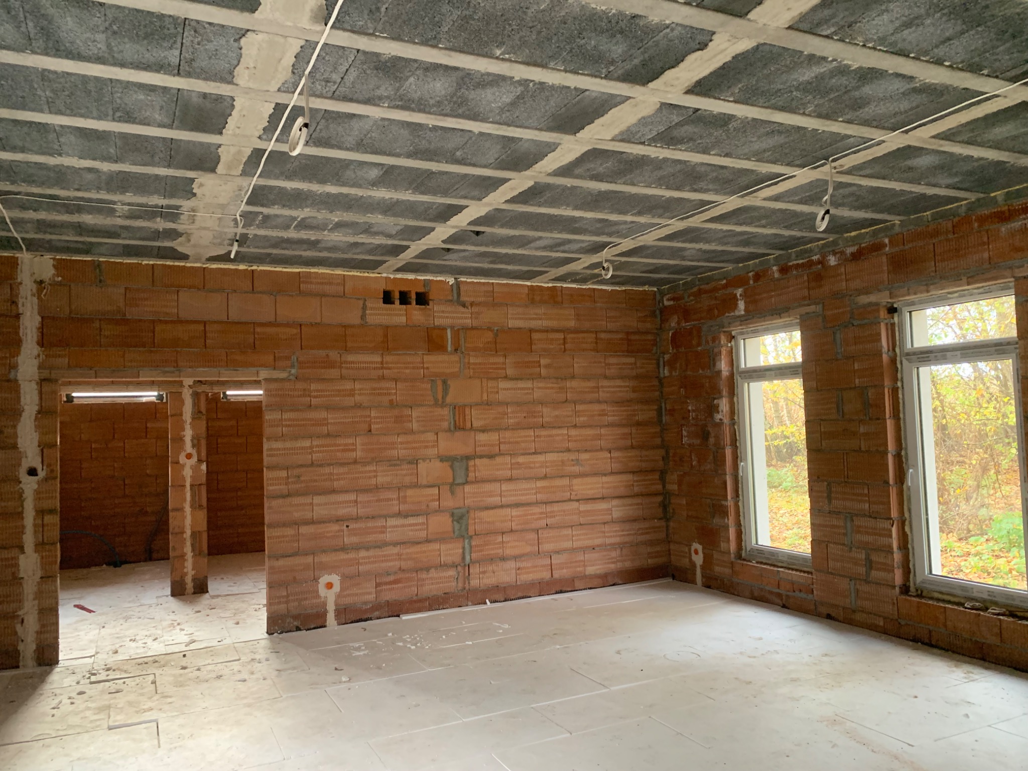 Surowy stan wnętrza domu z czerwonej cegły z oknami i betonowym stropem. Widoczne instalacje elektryczne. Podłoga wyłożona płytami izolacyjnymi.