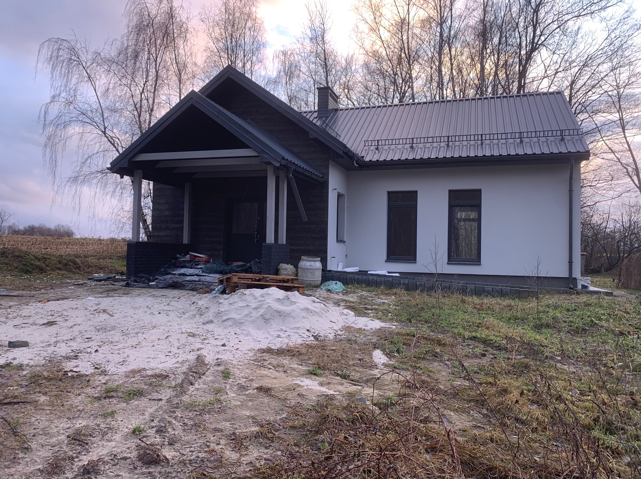 Nowoczesny dom z ciemną, zadaszoną werandą i jasną elewacją, otoczony ziemią i drzewami bez liści. Widoczny stos piasku przed budynkiem.