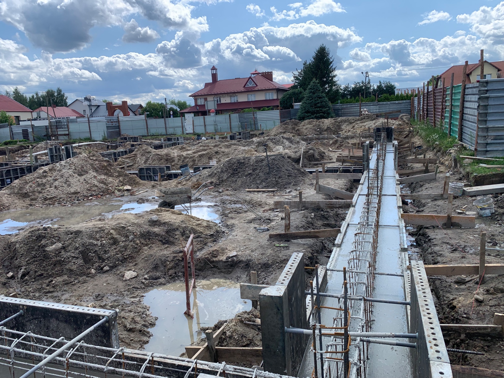 Wylane fundamenty z widocznym zbrojeniem na placu budowy domu jednorodzinnego w słoneczny dzień. W tle widoczne domy i błękitne niebo z chmurami.