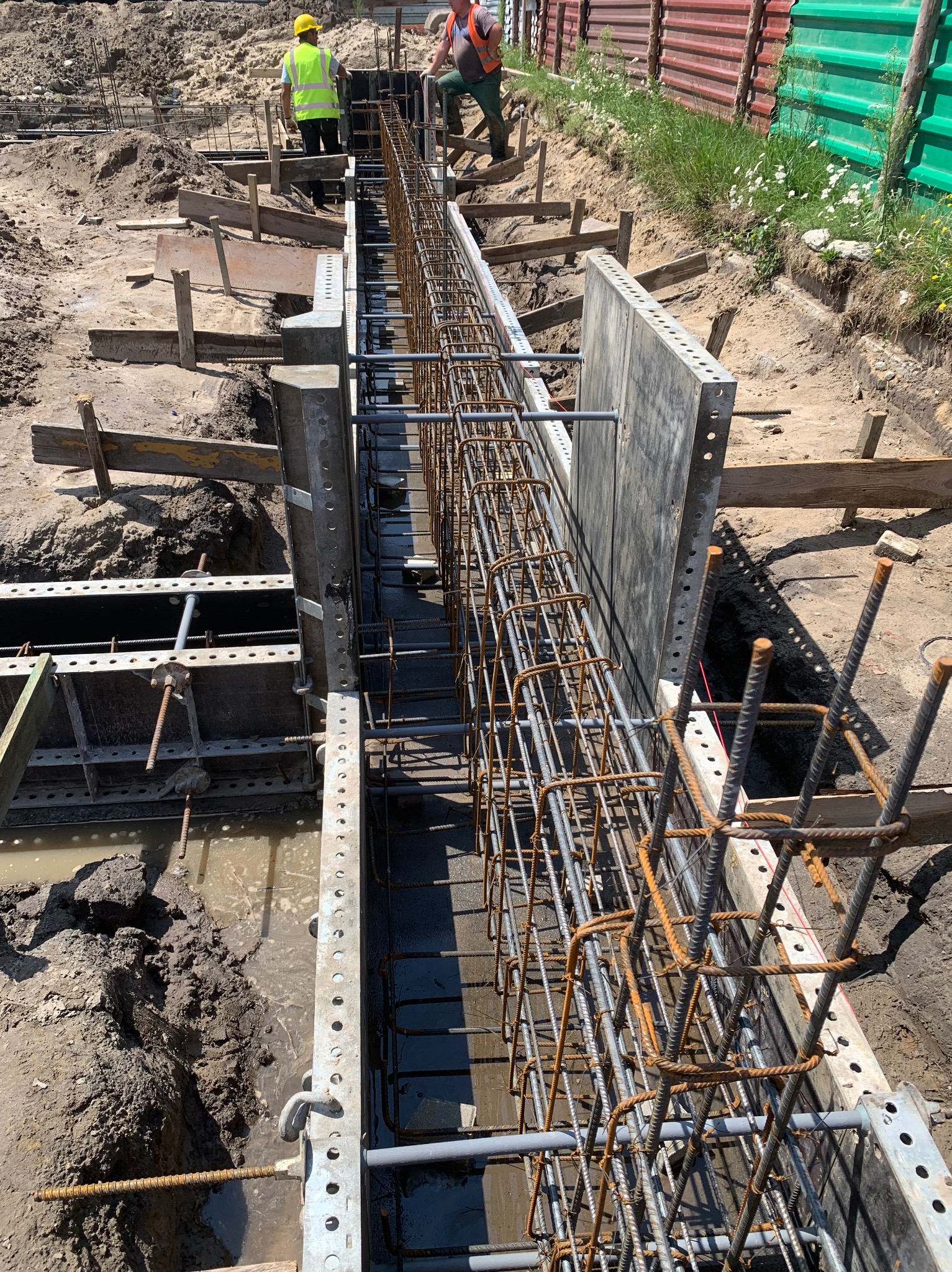 Zbrojenie w szalunku pod fundament, widoczni pracownicy w odblaskowych kamizelkach. Ujęcie z góry, ukazujące detale konstrukcji i przygotowania do zalewania betonem.
