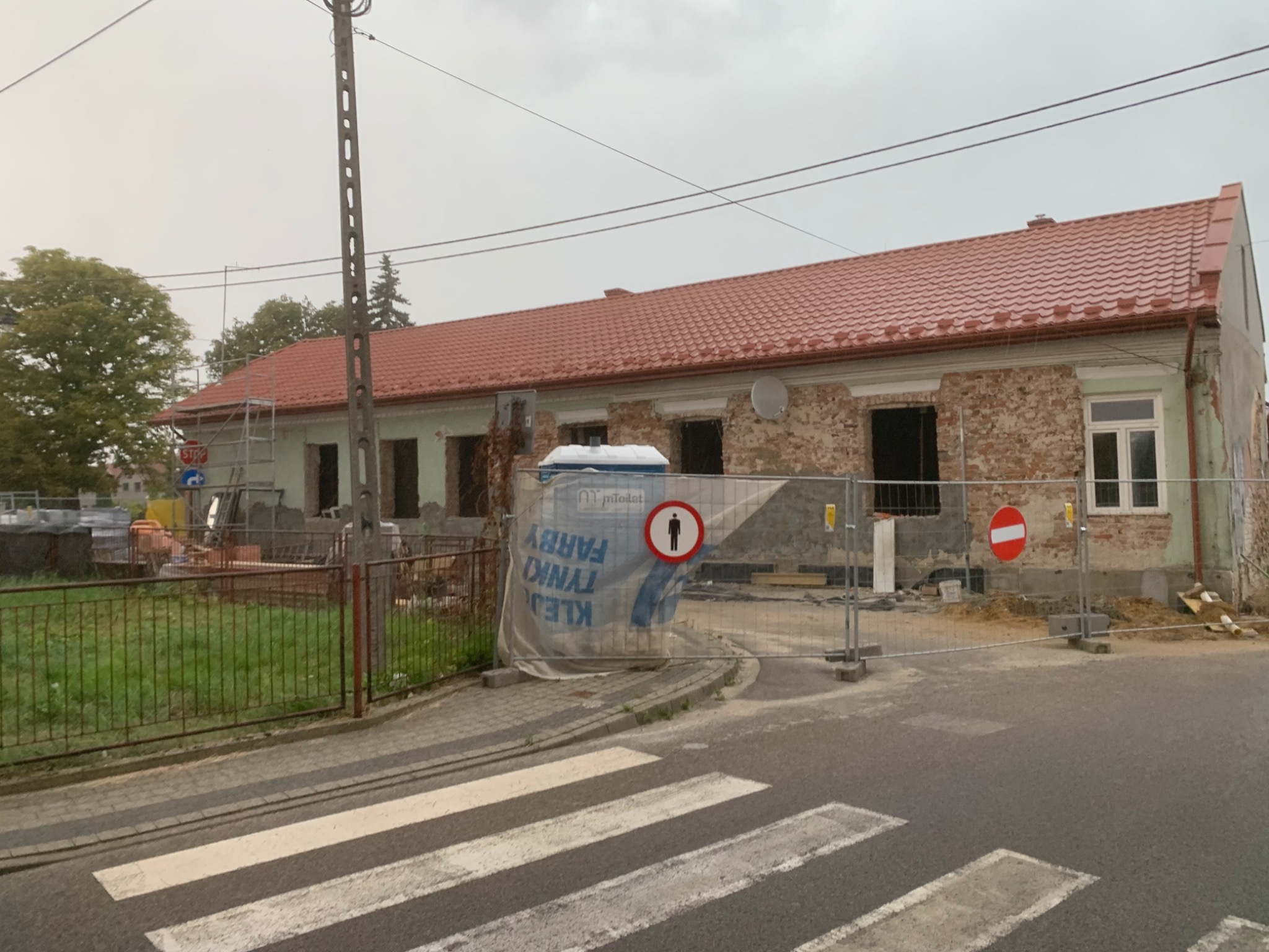 Budynek z czerwoną dachówką w trakcie remontu elewacji, zabezpieczony barierkami i siatką. Widoczne odsłonięte cegły i elementy rusztowania. Pochmurne niebo w tle.