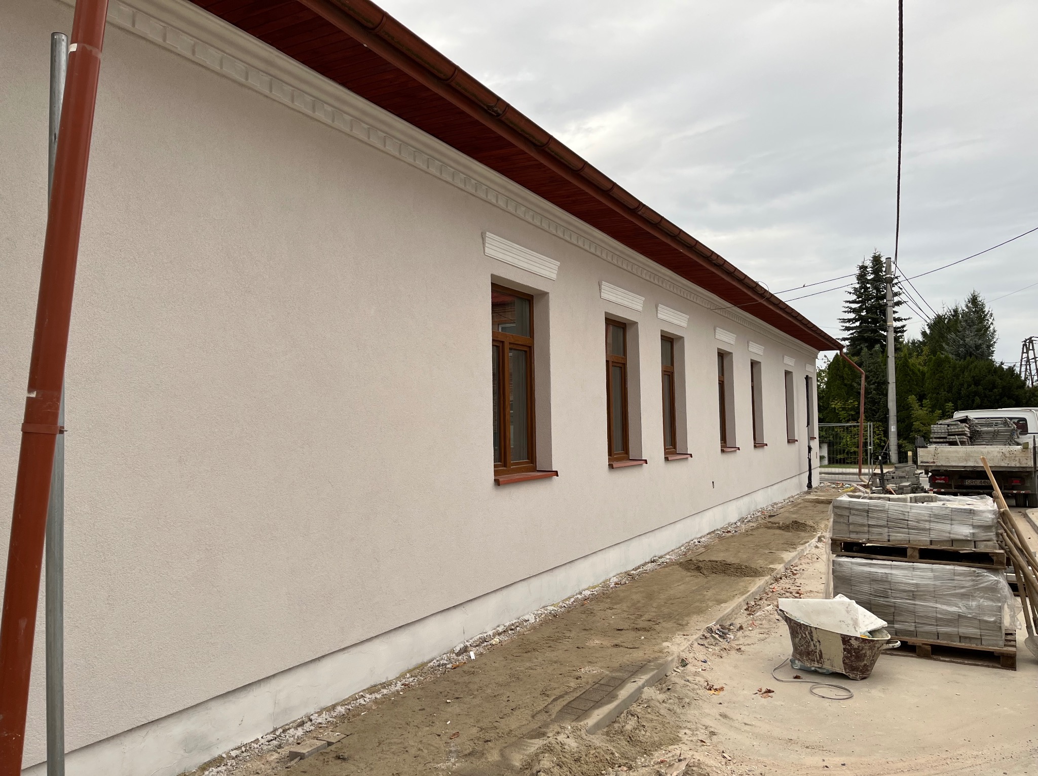 Ocieplony budynek z beżową elewacją, brązowymi oknami i czerwonym dachem. Widoczne palety z kostką brukową i narzędzia budowlane. Świeżo otynkowana ściana.