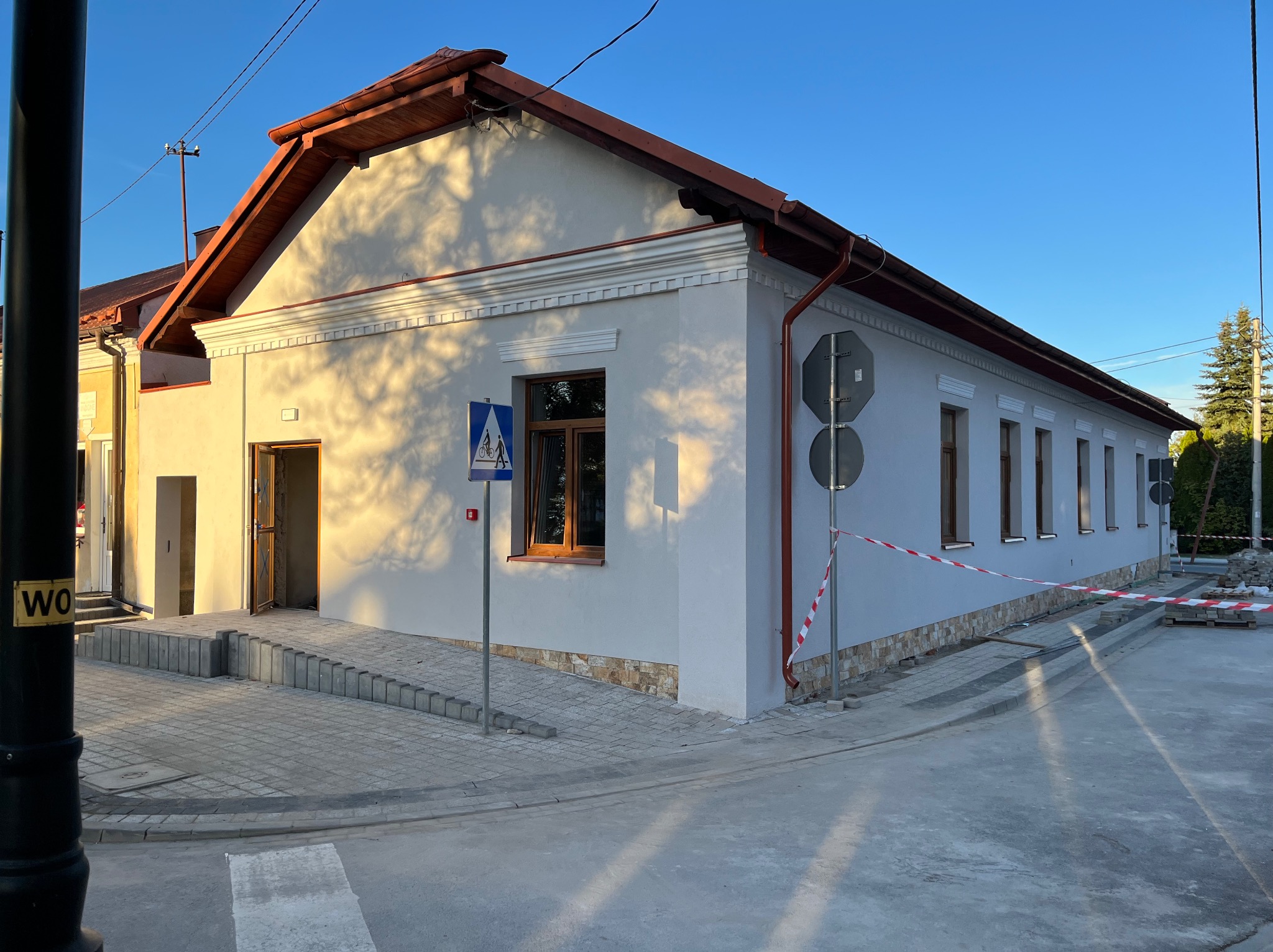 Odnowiony budynek z nową elewacją, drewnianą stolarką okienną i chodnikiem z kostki brukowej. Widoczny znak drogowy i taśma ostrzegawcza.