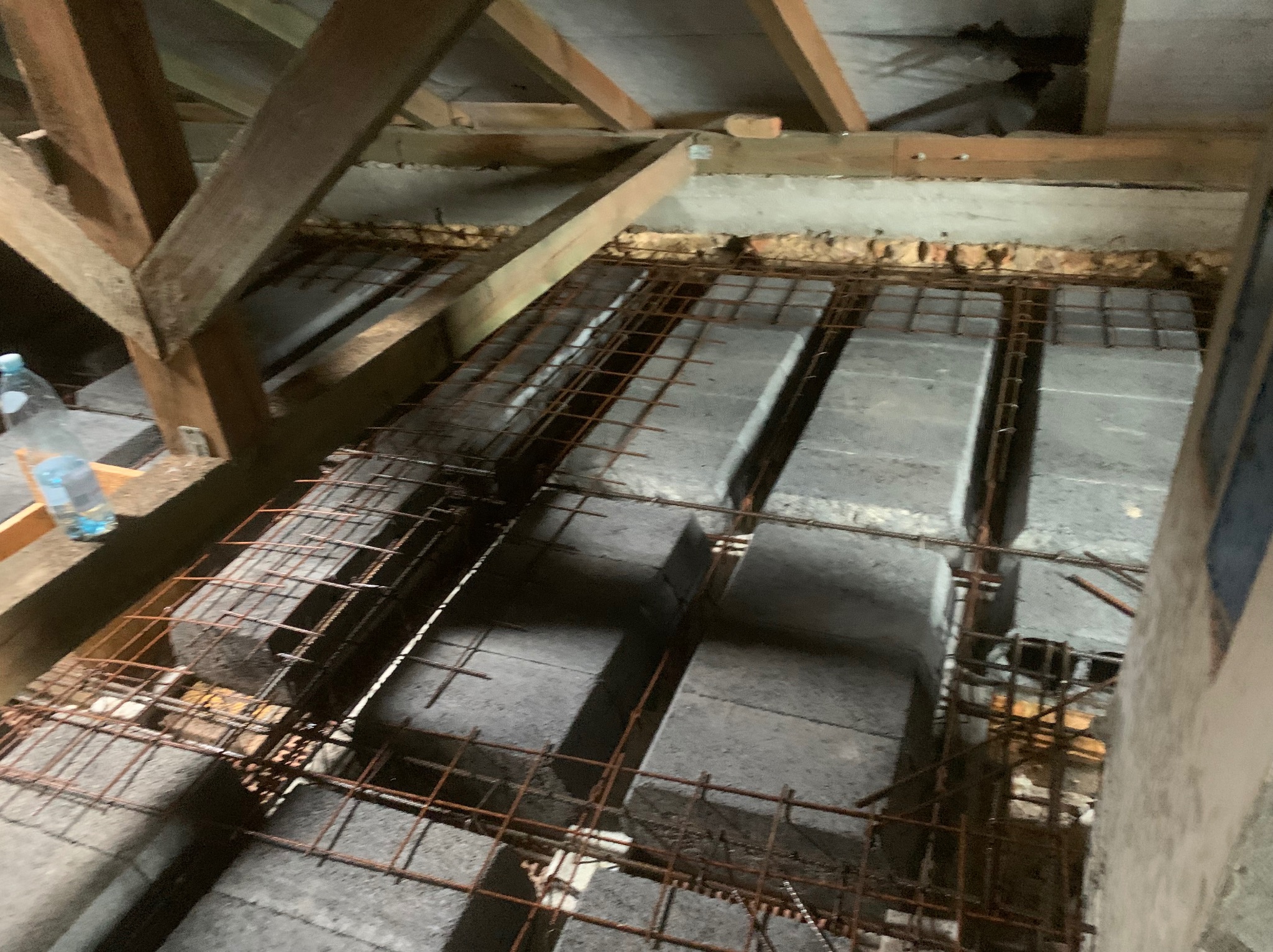 Zbrojenie stropu Teriva z betonowymi pustakami, widoczne drewniane elementy konstrukcji dachu i butelka wody. Ujęcie z góry, surowy stan budowy.