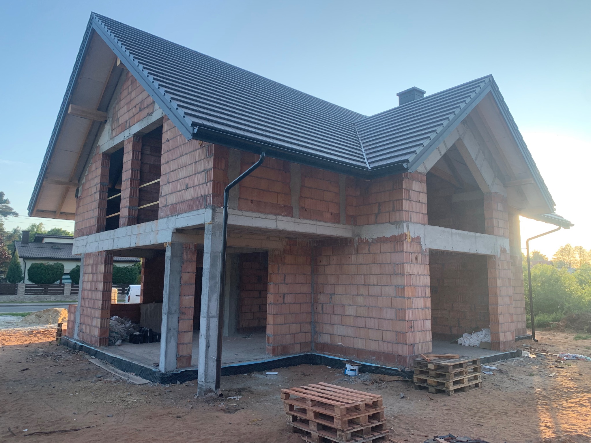 Surowy dom z czerwonej cegły z ciemnym dachem w trakcie budowy, widoczne palety. Dom w stanie deweloperskim, z zamontowanymi rynnami i oknami bez szyb.