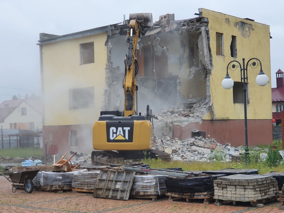 Żółta koparka CAT burzy budynek z żółtą elewacją. Wokół gruz i pył. Na pierwszym planie palety z kostką brukową i taczka. W tle widoczny fragment innego budynku.