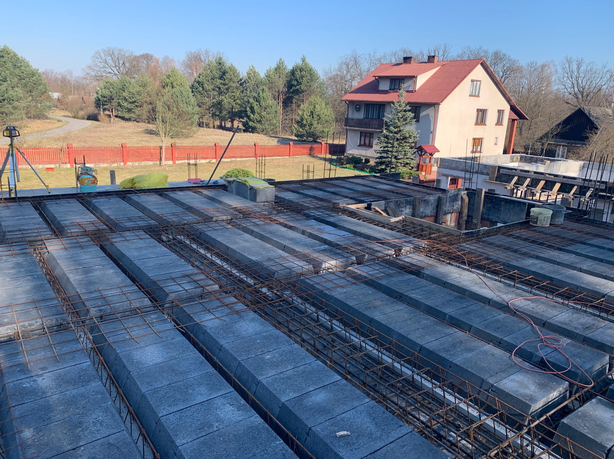 Budowa stropu Teriva z widocznym zbrojeniem i pustakami, w tle dom jednorodzinny i drzewa, słoneczny dzień.