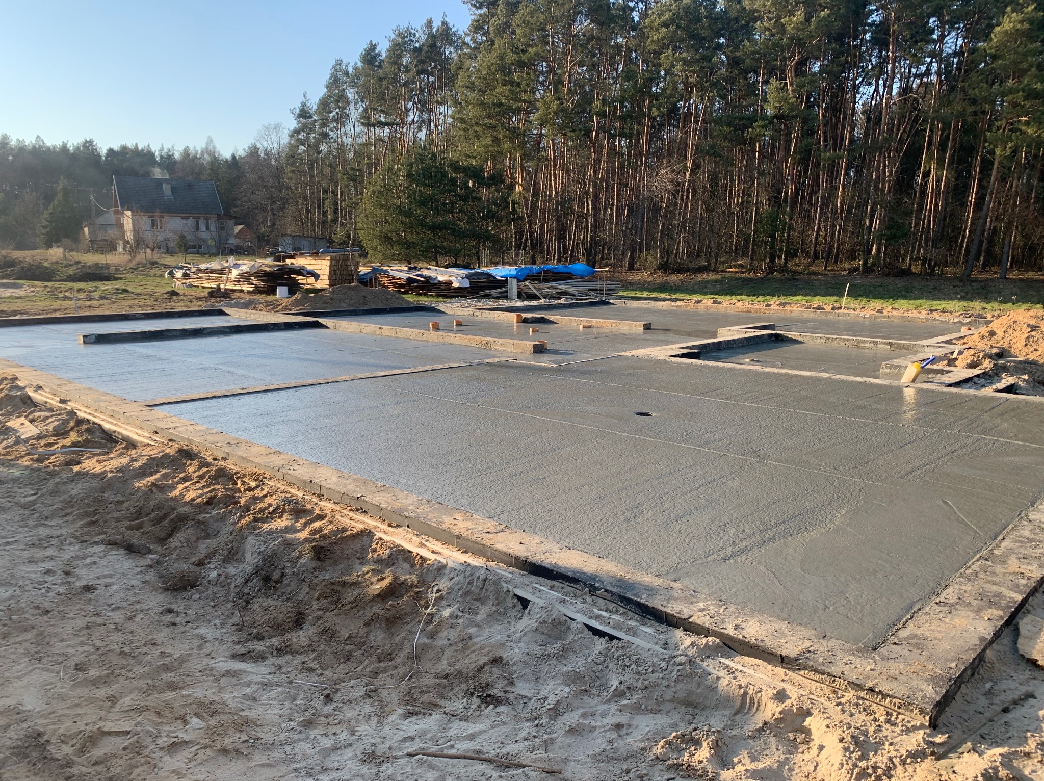 Świeżo wylany, gładki fundament pod budowę domu jednorodzinnego, otoczony piaskiem i zalesionym terenem. W tle widoczny budynek w stylu rustykalnym.