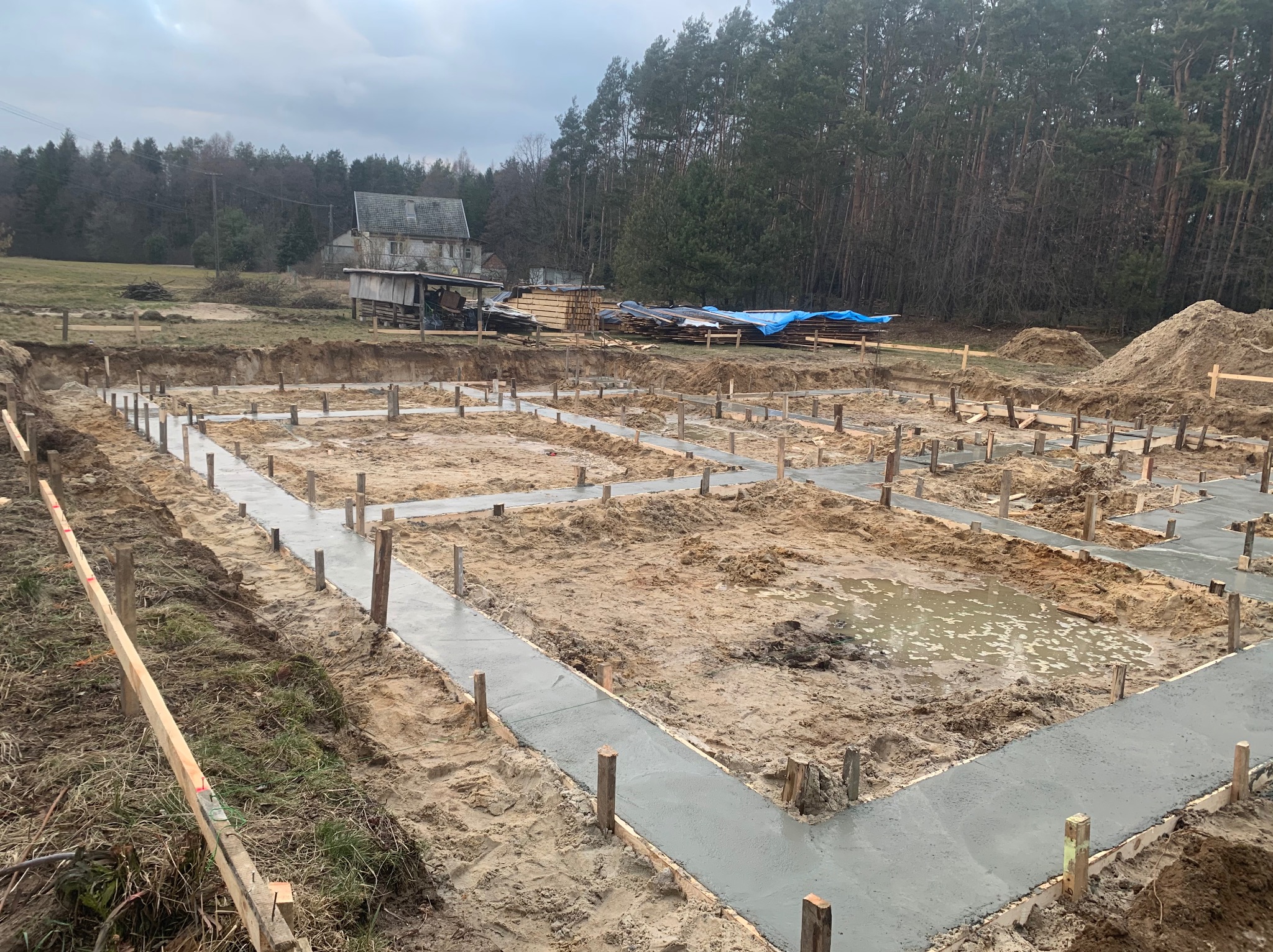 Wylane betonowe ławy fundamentowe z drewnianym szalunkiem na piaszczystym terenie budowy, w tle las i budynek gospodarczy. Widok z góry.