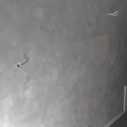 Ściana z efektem dekoracyjnym w odcieniach szarości, widoczny fragment instalacji elektrycznej i surowy brzeg ściany przy futrynie, fragment schodów z drewnianą barierką.