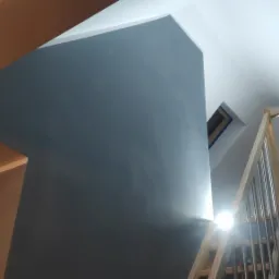 Świeżo pomalowana szara ściana w przestrzeni klatki schodowej z drewnianymi schodami z metalowymi poręczami, widok z dołu, światło słoneczne wpadające przez okno dachowe.