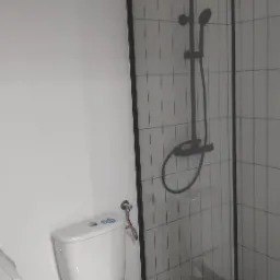 Nowoczesna łazienka z białą toaletą i kabiną prysznicową z czarnymi ramami oraz deszczownicą na tle białych płytek.