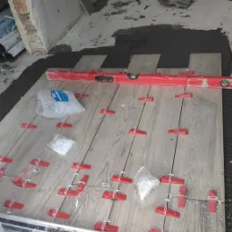 Układanie płytek imitujących drewno na podłodze, widoczny poziomica, krzyżyki dystansowe i woreczki z klinami, w tle osoba w ubraniu roboczym.