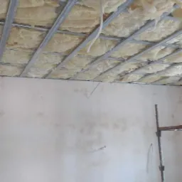 Montaż izolacji z wełny mineralnej w metalowym stelażu sufitu podwieszanego, widoczne przewody elektryczne i fragment ściany z instalacją elektryczną, drabina i rusztowanie w tle.