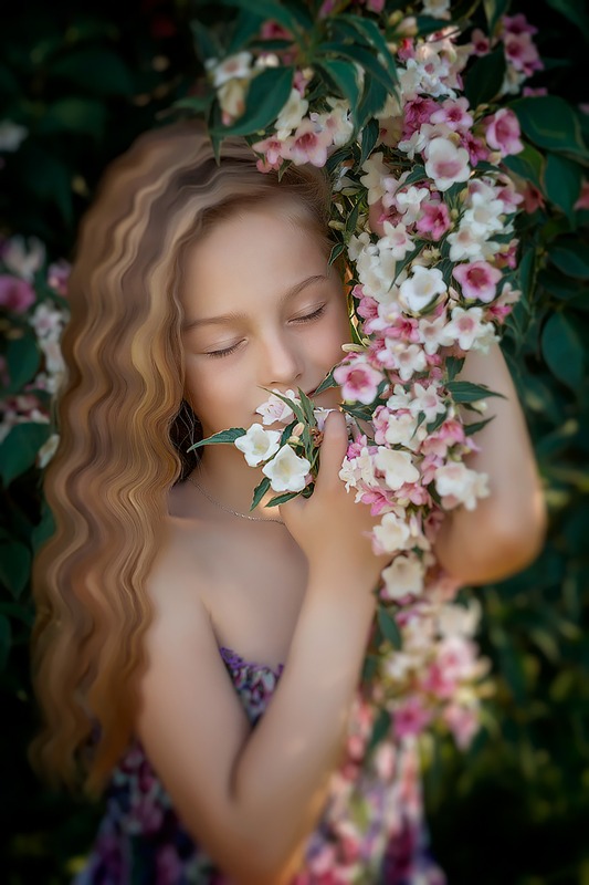 Dziewczynka z długimi blond włosami, z zamkniętymi oczami, wącha gałązkę kwitnącego krzewu o białych i różowych kwiatach.