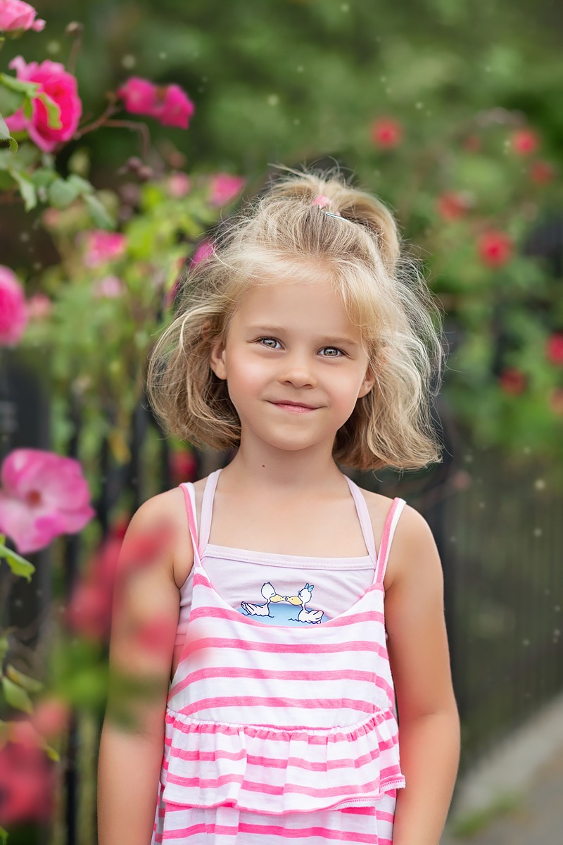 Portret uśmiechniętej blond dziewczynki w różowo-białej sukience na tle kwitnących różowych róż i zieleni.