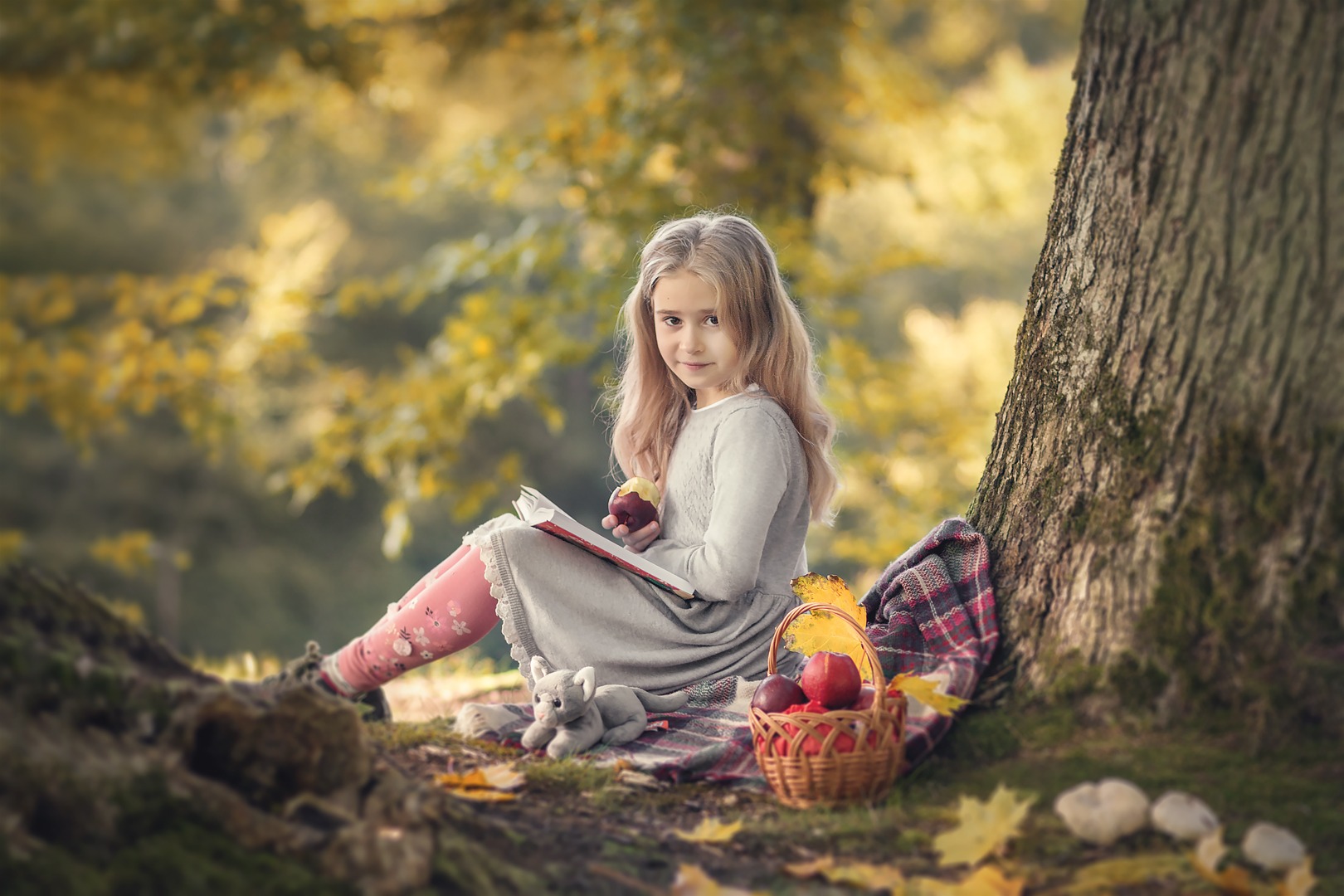 Dziewczynka w szarej sukience siedzi pod drzewem, czyta książkę i trzyma czerwone jabłko, obok koszyk z owocami i pluszowy kotek, jesienne liście w tle.
