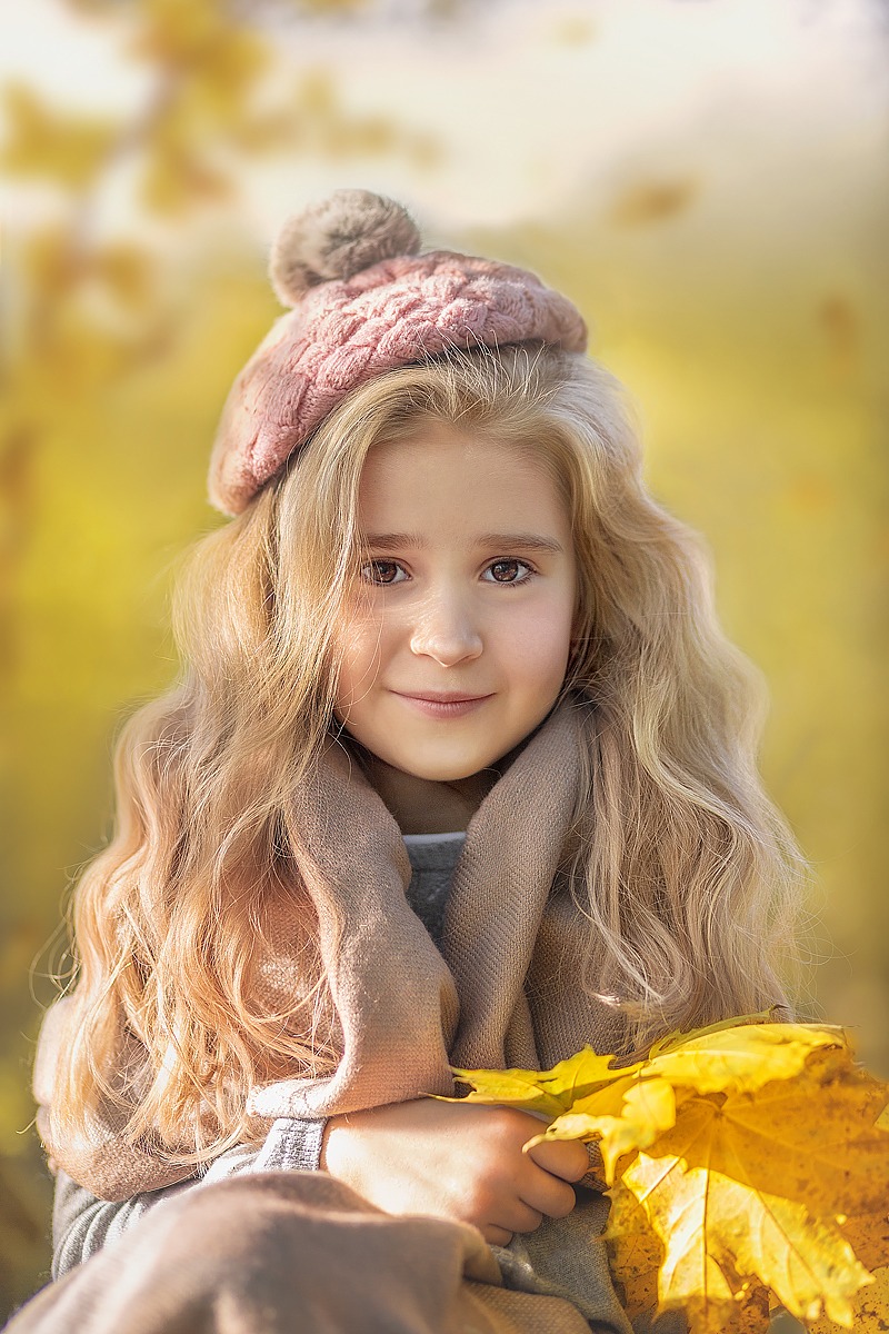 Portret uśmiechniętej dziewczynki z blond lokami w różowej czapce z pomponem, otulonej beżowym szalem, trzymającej w rękach żółte liście klonu na rozmytym, jesiennym tle.