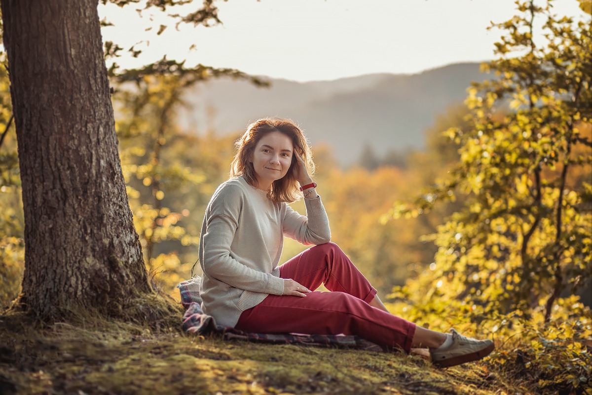Kobieta siedząca pod drzewem w jesiennym lesie, opierająca głowę o dłoń, z uśmiechem spogląda w obiektyw, w tle rozmyte wzgórza.