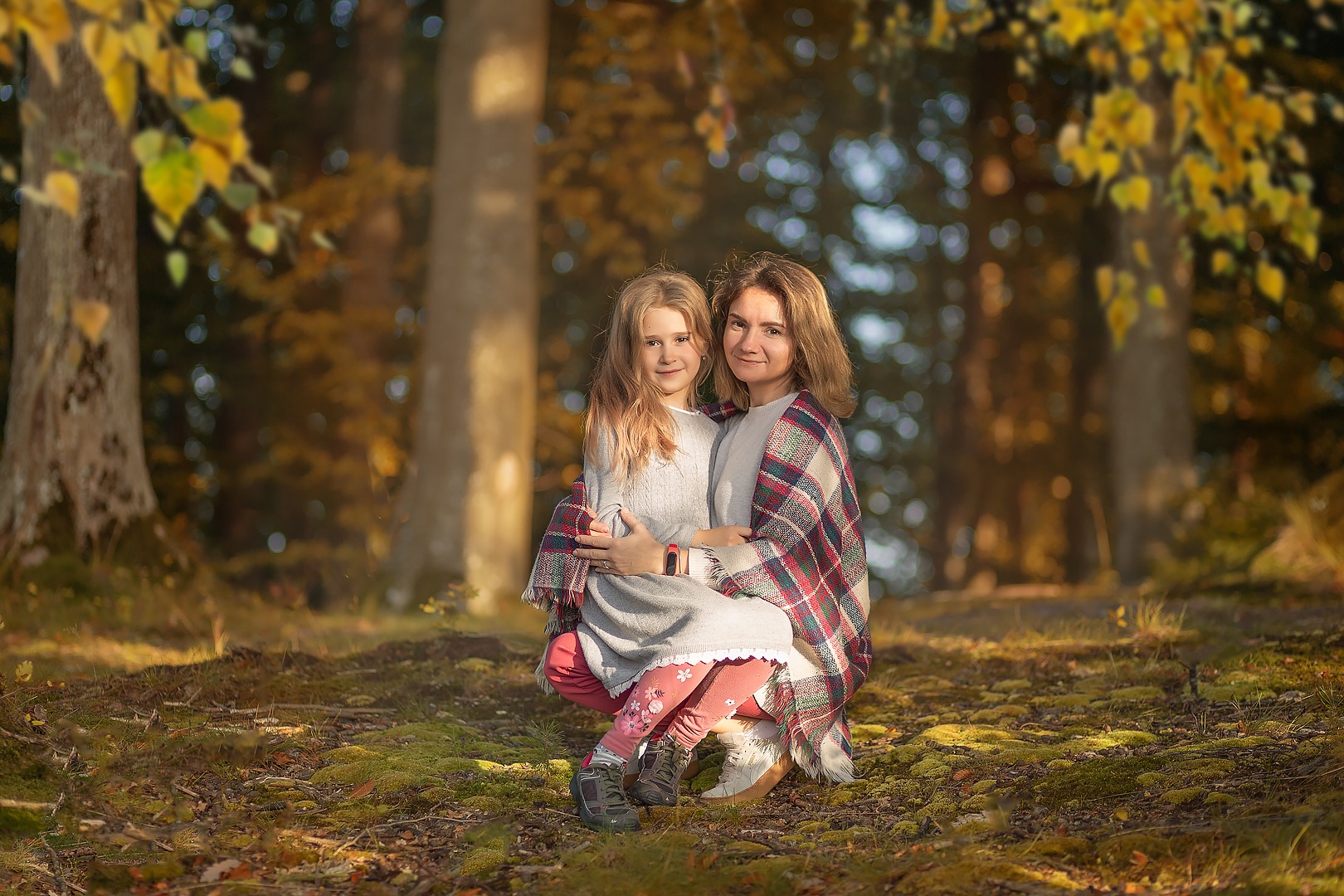 Matka i córka tulą się do siebie w jesiennym lesie, okryte pledem w kratę, siedząc na mchu w promieniach popołudniowego słońca.