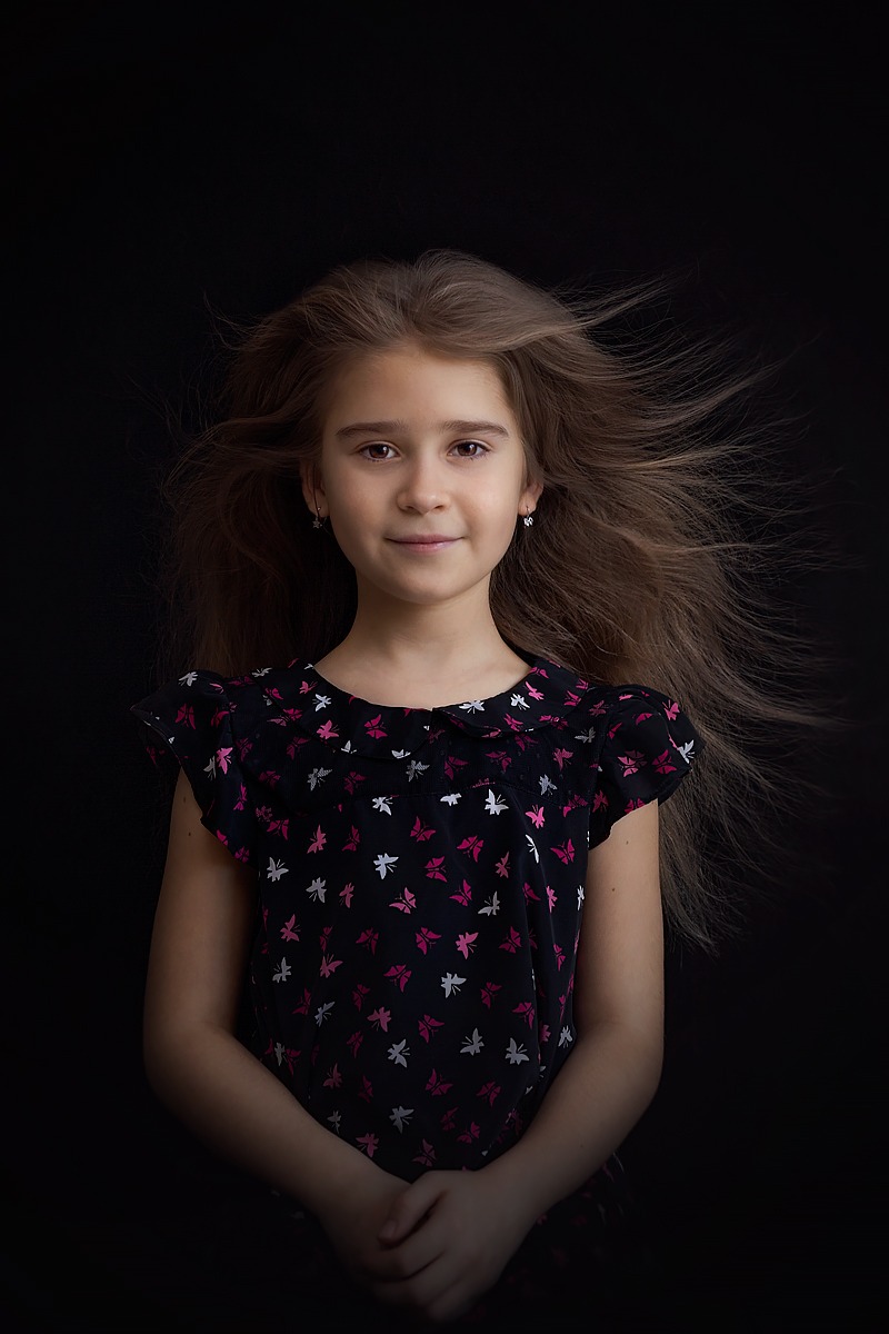 Portret uśmiechniętej dziewczynki z rozpuszczonymi włosami na czarnym tle, ubrana w bluzkę w motyle.