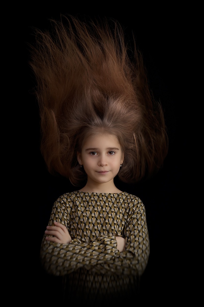 Portret dziewczynki z rozwichrzonymi, uniesionymi włosami na czarnym tle, ubrana w bluzkę z geometrycznym wzorem, ręce skrzyżowane.