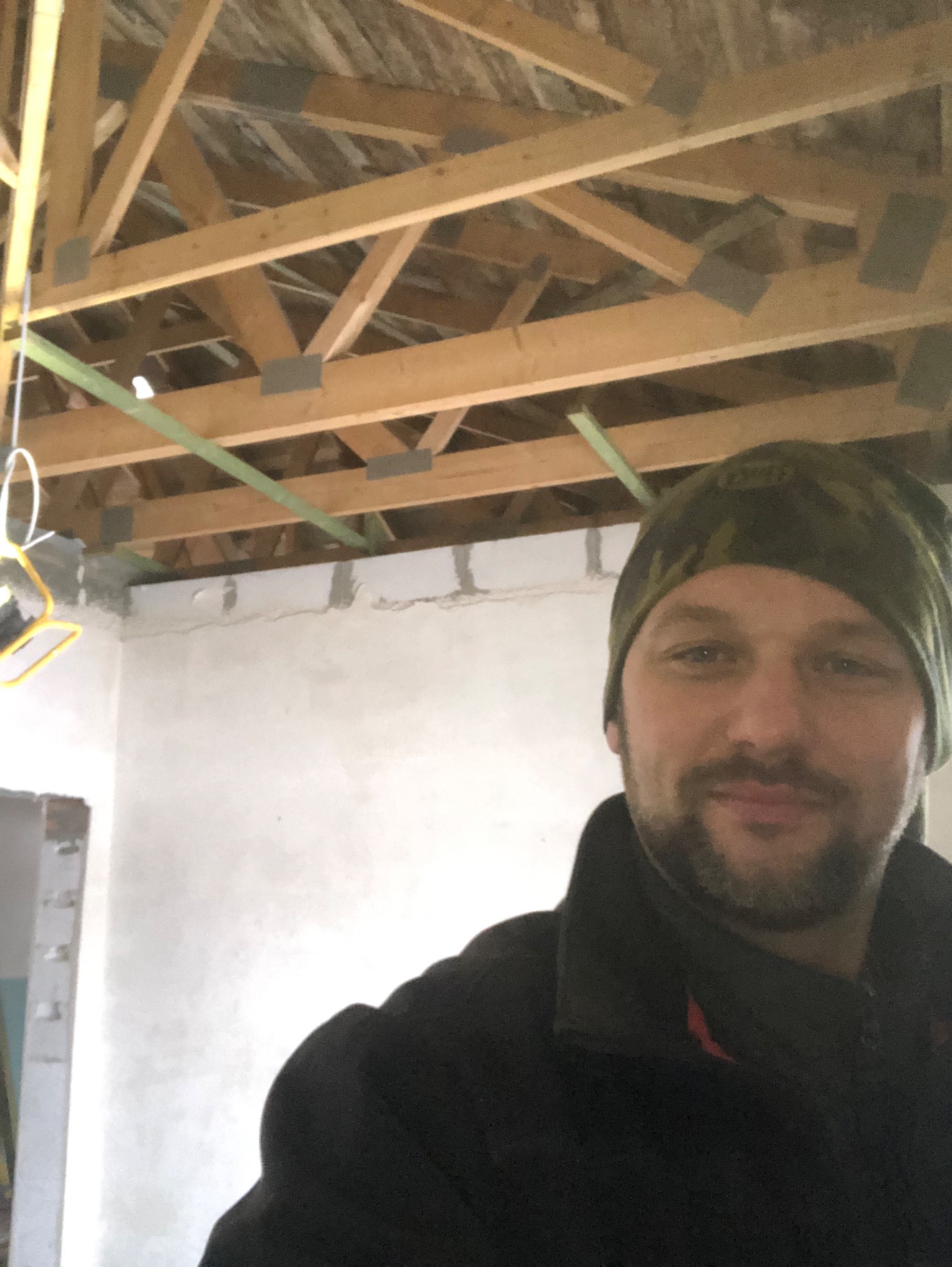 Mężczyzna w czapce na tle surowego stanu poddasza z widoczną konstrukcją drewnianą dachu i fragmentem otynkowanej ściany.