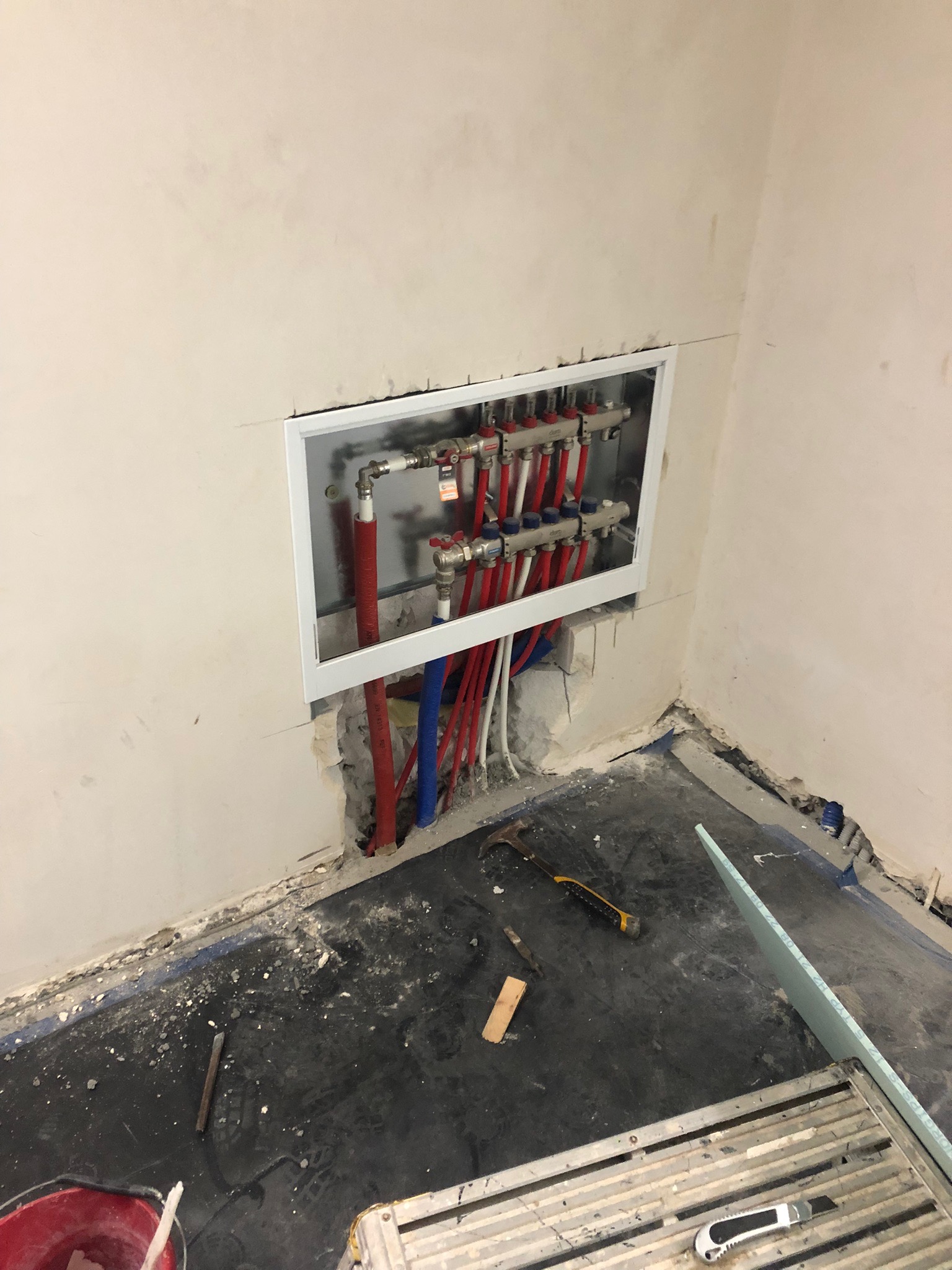 Rozdzielacz instalacji centralnego ogrzewania wnękowy, z czerwonymi i niebieskimi rurami, widoczny w trakcie montażu w surowym wnętrzu, z gruzem na podłodze.