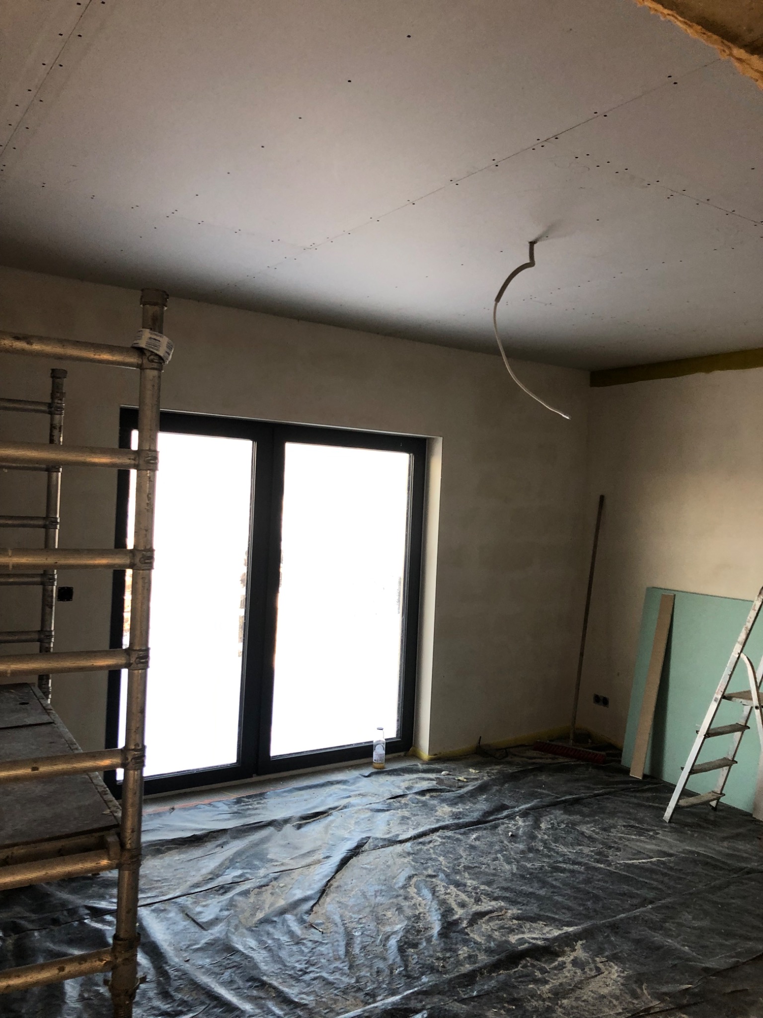 Wnętrze w trakcie prac wykończeniowych, widoczny sufit z zamontowaną płytą kartonowo-gipsową, zabezpieczona folią podłoga, rusztowanie i drabina.