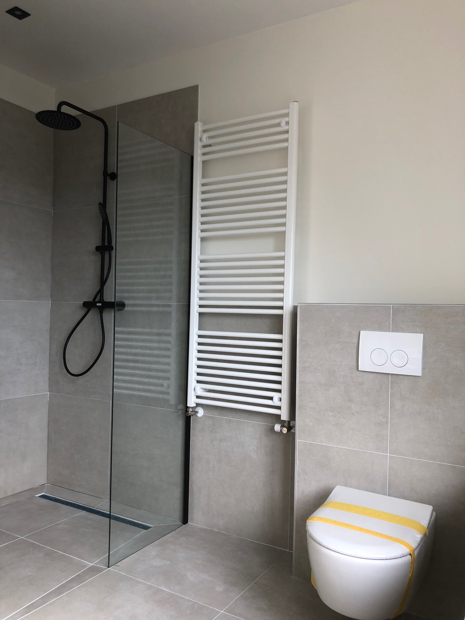 Nowoczesna łazienka po remoncie: kabina prysznicowa typu walk-in z czarną armaturą, biały grzejnik drabinkowy i podwieszana toaleta zabezpieczona folią, szare płytki na ścianach i podłodze.
