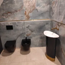 Wykończona łazienka z marmurowymi ścianami w odcieniach szarości i beżu, czarną armaturą i złotymi akcentami, prezentująca nowoczesny design z wiszącą toaletą i bidetem.