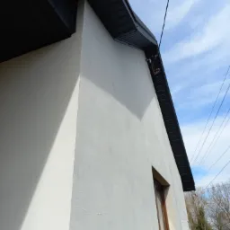 Fragment narożnika budynku z nową, ciemną podbitką dachową i widocznymi przewodami na tle błękitnego nieba z chmurami.