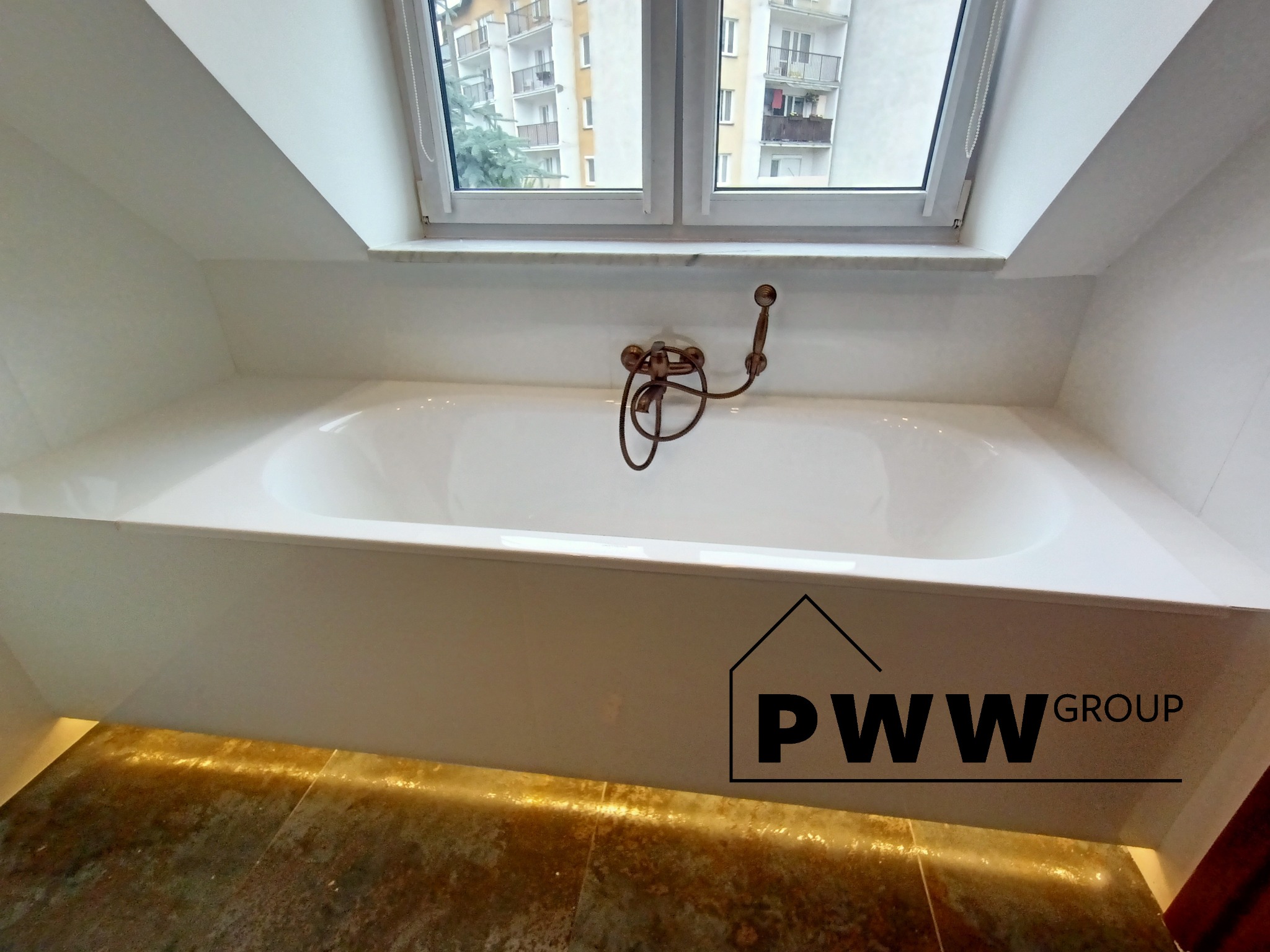 Biała wanna z mosiężną armaturą umieszczona pod oknem na poddaszu, z podświetleniem LED u dołu obudowy wanny. Widoczne logo firmy na obudowie wanny.