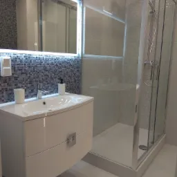 Nowoczesna łazienka z umywalką na szafce, kabiną prysznicową ze szklanymi drzwiami i mozaiką na ścianach, oświetlona lustrem.