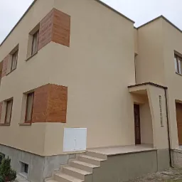 Nowoczesny dom jednorodzinny z beżową elewacją i panelami dekoracyjnymi imitującymi drewno, widoczne schody wejściowe i biała skrzynka na media.