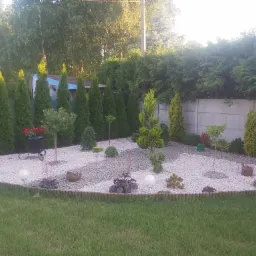 Ogród z białym żwirem, ozdobnymi drzewkami i rabatami, obrzeżony drewnianymi palisadami, z tujami w tle i betonowym ogrodzeniem.